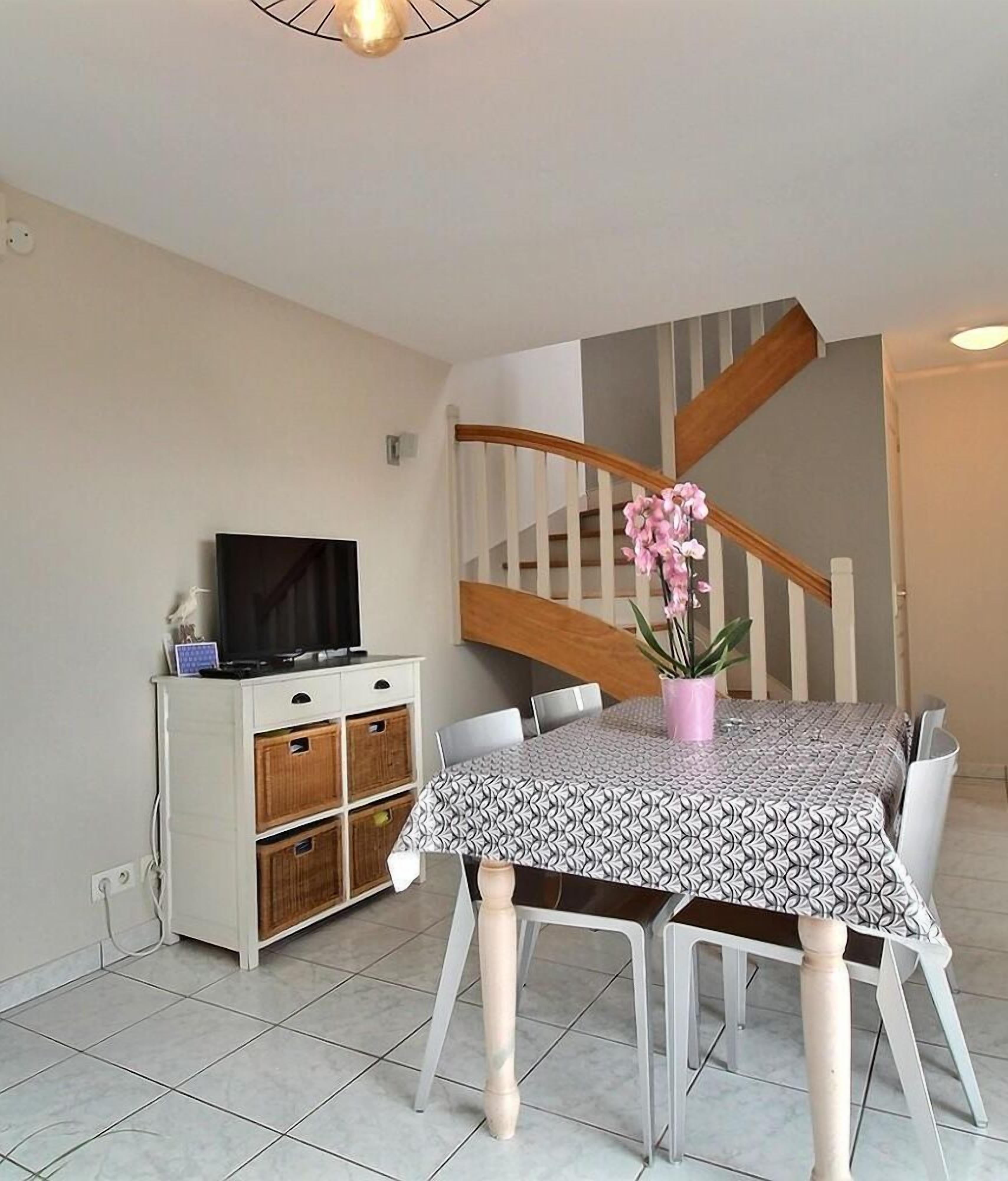 A dining area with a table set for four, a television on a cabinet, and a staircase in the background.