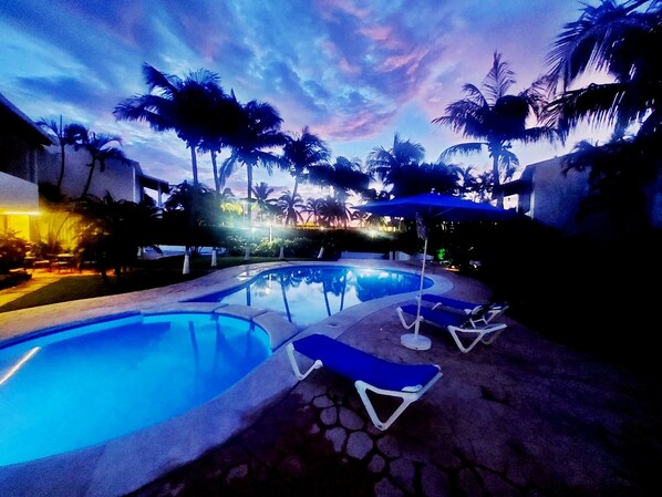 A swimming pool with lounge chairs and palm trees, surrounded by buildings.
