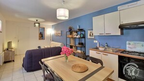 A compact kitchen with a stove, oven, and white cabinets. A dining table with chairs is positioned in the center, with a vase of flowers on it. - Le Grand Kergroas - LANNEBERT (LANNEBERT)