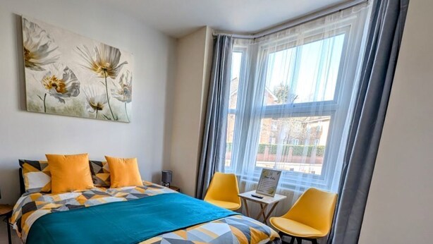 Une chambre d’hôtel avec un lit, deux chaises, une petite table, une fenêtre avec des rideaux et un tableau au mur.