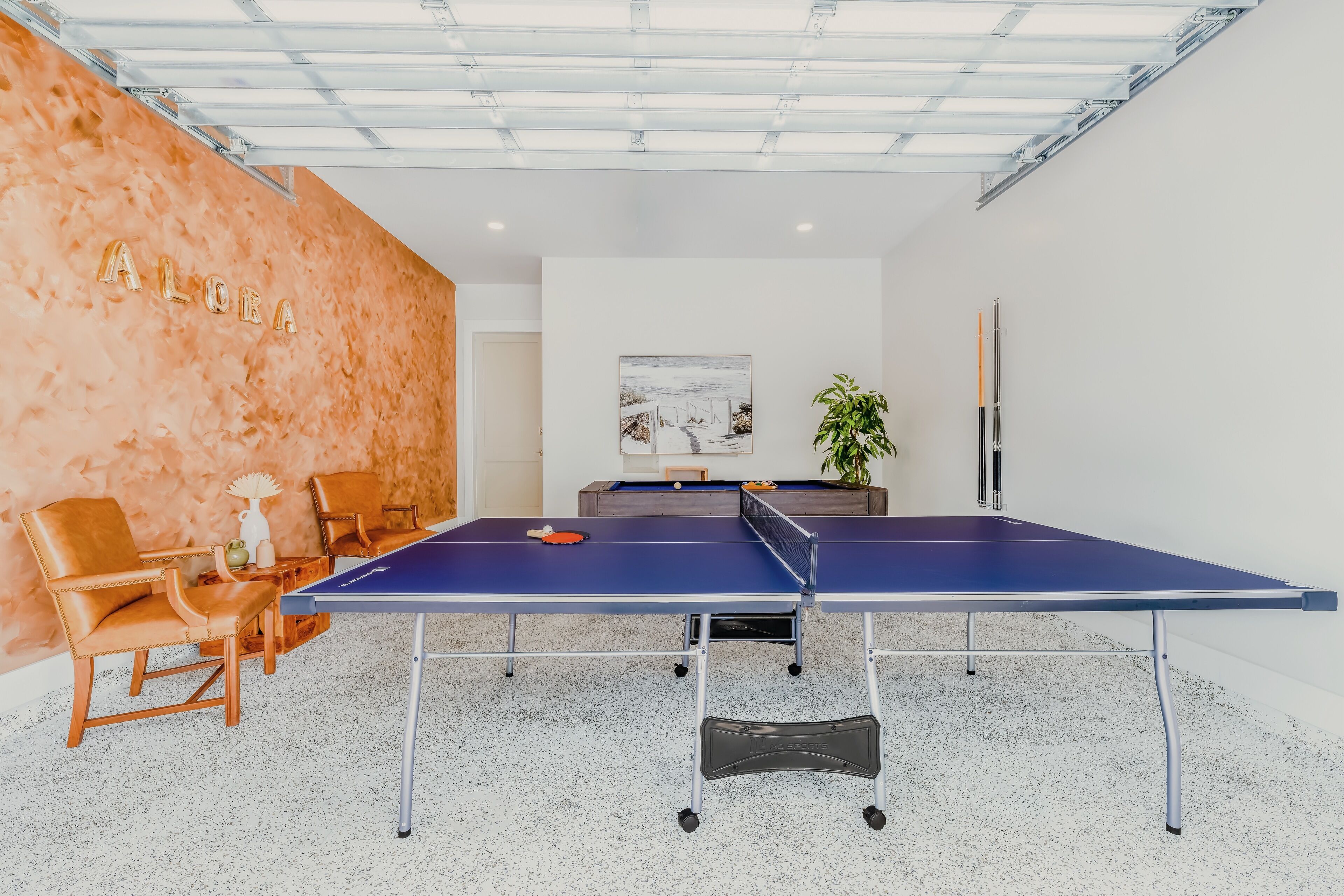 A ping pong table in a room with a textured orange wall, a painting, and a plant.
