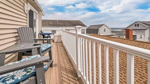 A wooden deck with a white railing, a table with chairs, and a view of neighboring houses and the ocean.