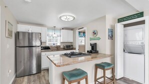 A modern kitchen with white cabinets, a stainless steel refrigerator, a built-in microwave, and a coffee machine. There are two bar stools with teal cushions.