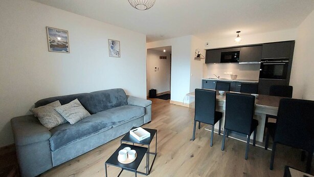 A modern living room with a grey sofa, a coffee table, and a dining area with chairs and a kitchen in the background. - Cozy apartment 6 pers, ski-in access, parking included (Valmorel)