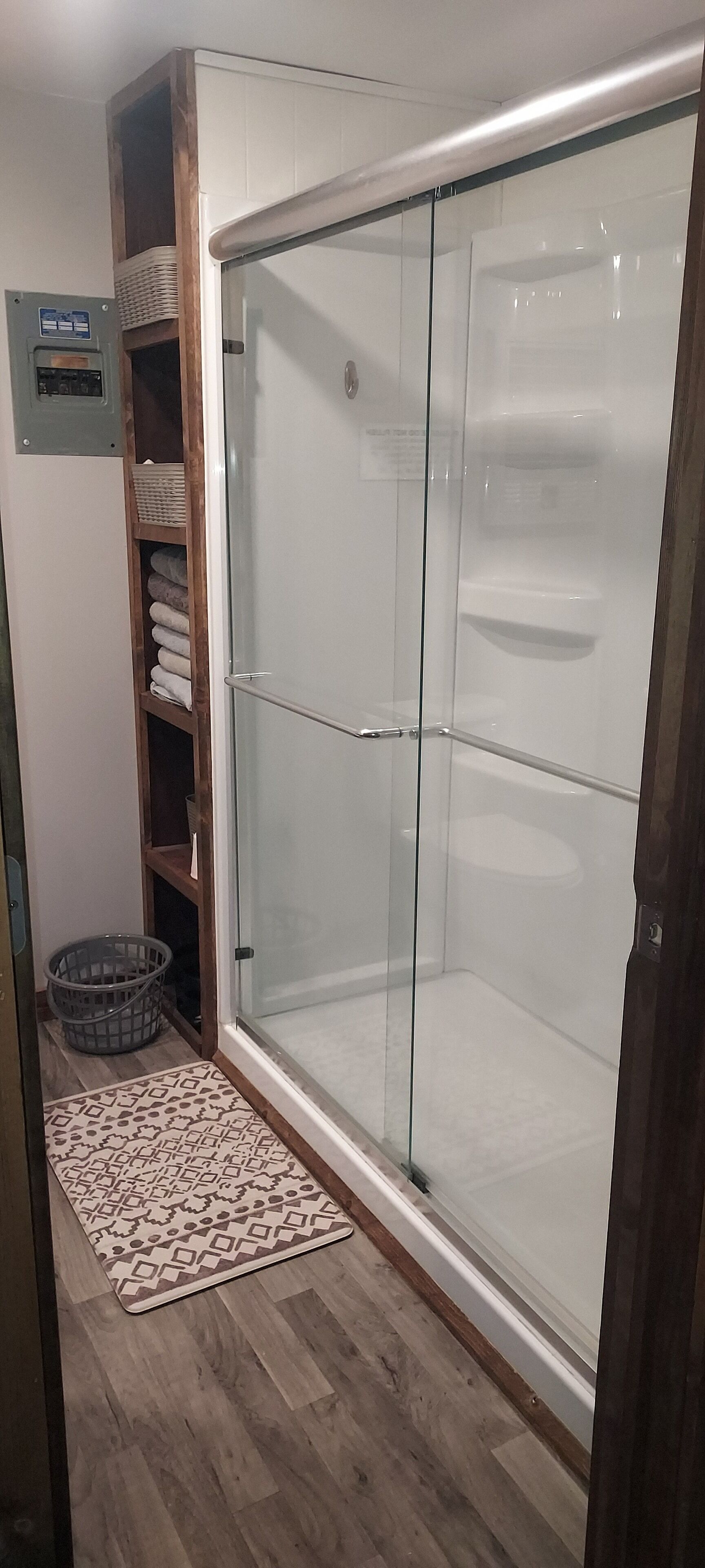 Una ducha a nivel con puertas de vidrio, un estante de madera con toallas dobladas y un tapete estampado en el suelo.