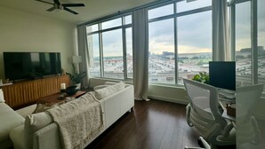 A modern living room with a large window, a ceiling fan, a flat-screen TV, a white sofa, a desk with a chair, and a view of a cityscape. - Stylish, centrally-located condo in Uptown/West Village with high-end amenities! (Dallas)