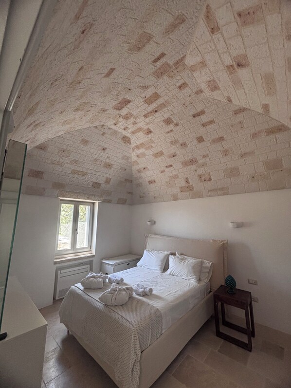 A bedroom with a brick-patterned ceiling, a large bed with white linens, a bedside table, and a window with a view of greenery.