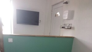 Une pièce comprenant un lit, un téléviseur à écran plat fixé au mur, une porte avec une fenêtre et une petite étagère sur laquelle sont posés divers objets.