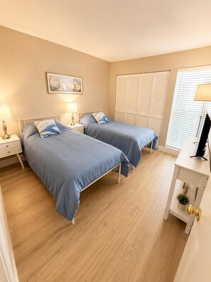 A bedroom with two beds, a bedside table with lamps, a window with blinds, and a desk with a computer. - Charming 2 BR condo with golf course views in North Tampa! Snowbirds welcome! (Tampa)