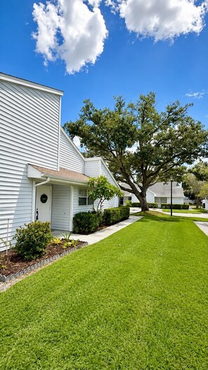 A residential area with a well-maintained lawn, a large tree, and houses with white siding. - Charming 2 BR condo with golf course views in North Tampa! Snowbirds welcome! (Tampa)