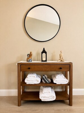 A wooden vanity with a round mirror, two decorative items, and three shelves holding folded towels. - Charming 2 BR condo with golf course views in North Tampa! Snowbirds welcome! (Tampa)
