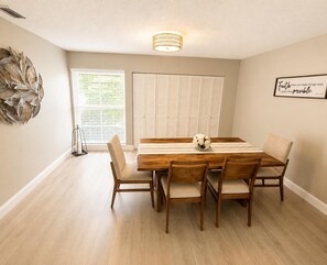 A dining room with a wooden table and chairs, a decorative wall piece, and a window with blinds. - Charming 2 BR condo with golf course views in North Tampa! Snowbirds welcome! (Tampa)