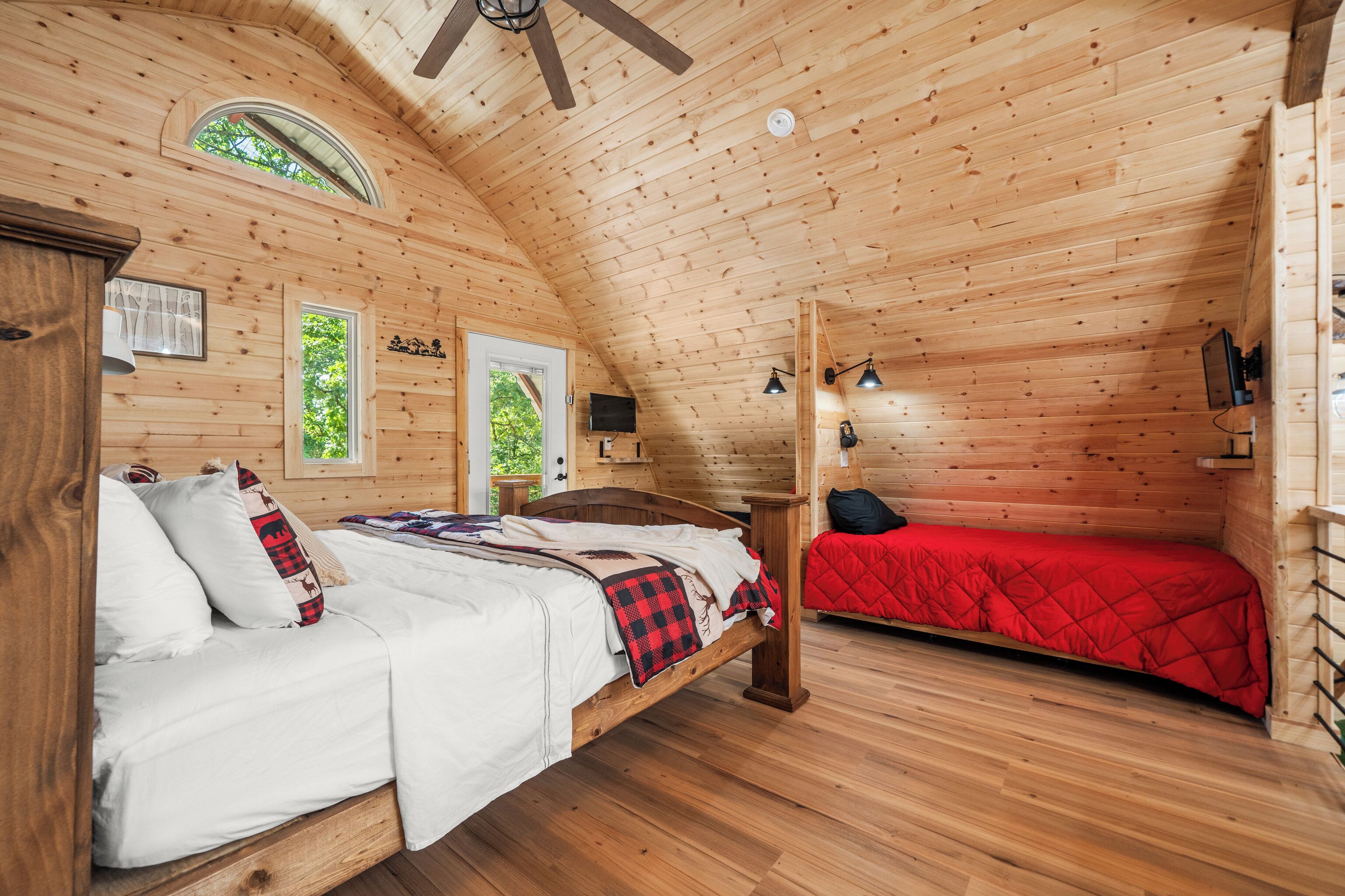 Una cabaña de madera con un dormitorio estilo altillo, una cama con ropa blanca y un colcha roja, y una pequeña zona de escritorio con una silla.