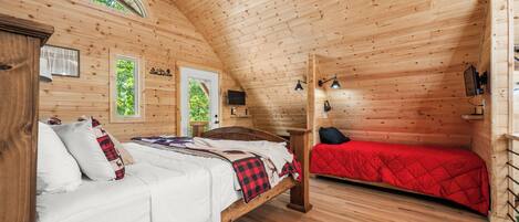 Una cabaña de madera con un dormitorio estilo altillo, una cama con ropa blanca y un colcha roja, y una pequeña zona de escritorio con una silla.