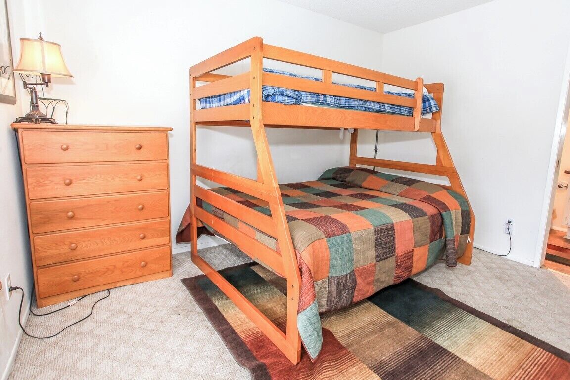 Een stapelbed met een geruit dekbed, een houten dressoir met lades, en een lamp op het dressoir.