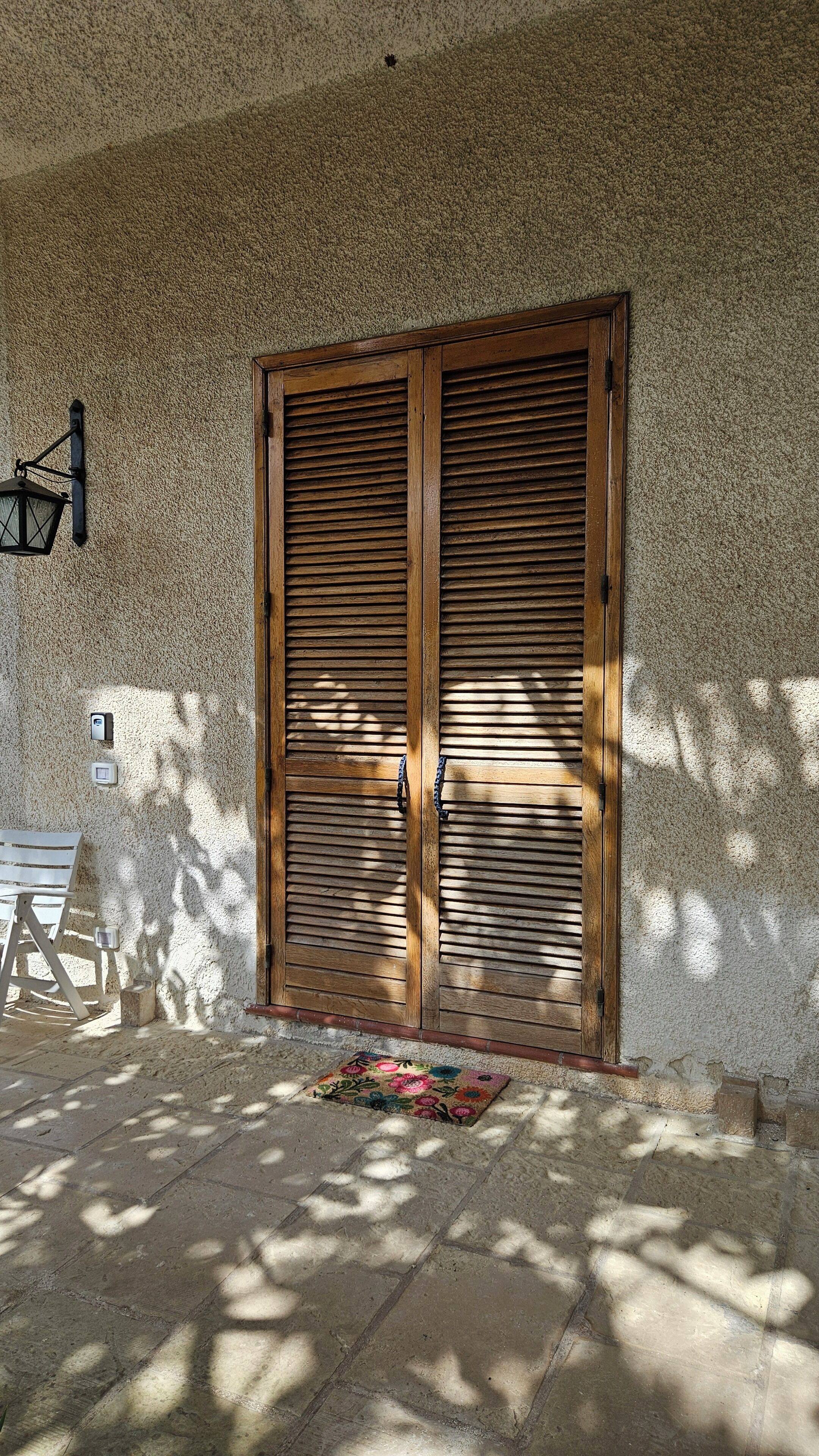A wooden door with horizontal slats, a wall-mounted light fixture, and a doormat on a stone-paved entrance.