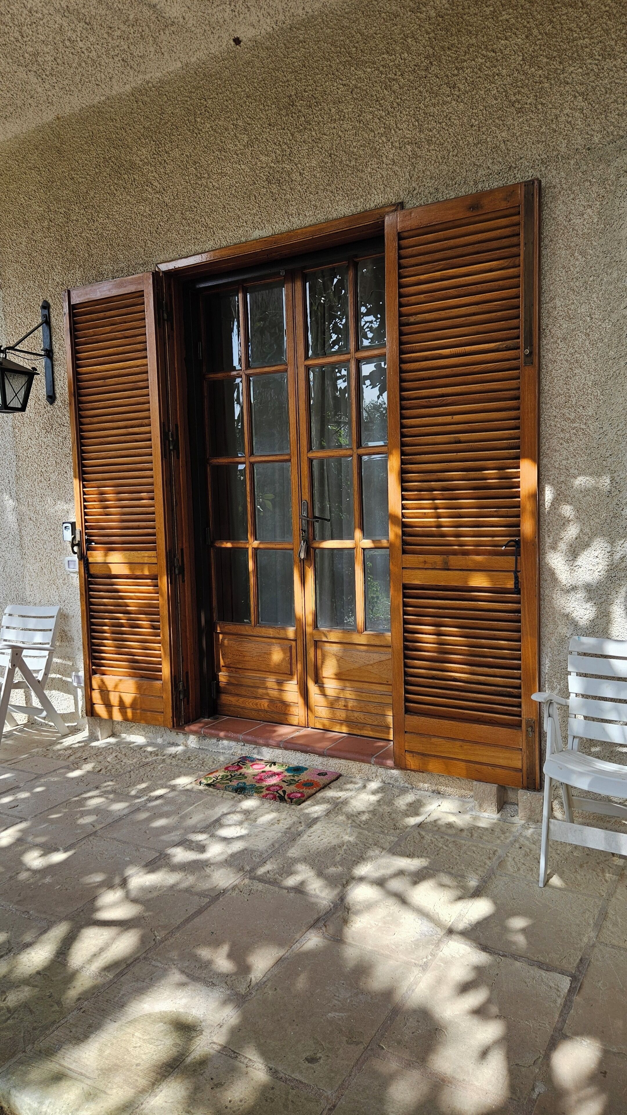Una puerta de madera con un panel de vidrio, flanqueada por contraventanas de madera, que da a un acceso empedrado.