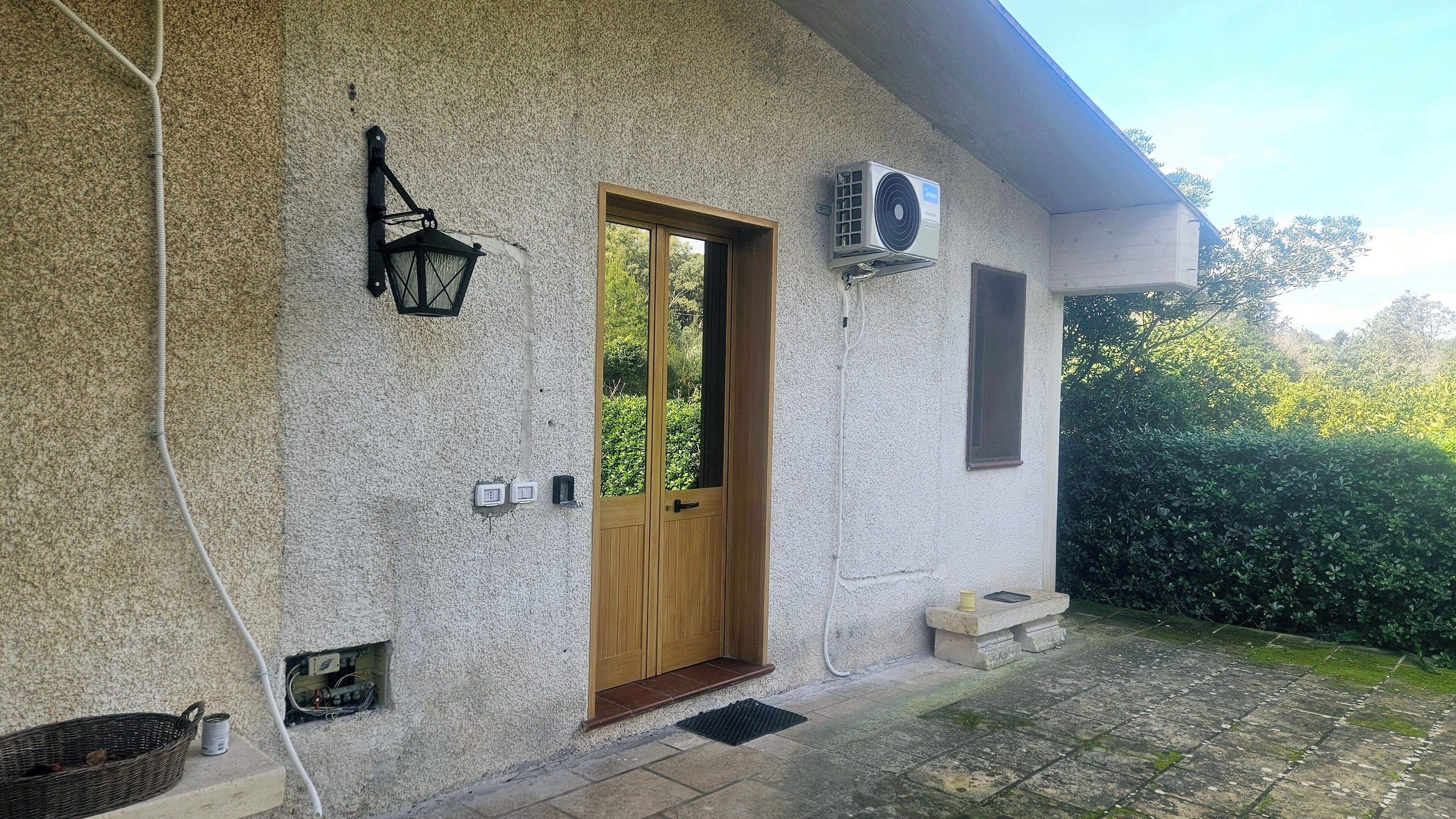 Un bâtiment à l’extérieur texturé, avec une porte en bois, un climatiseur fixé au mur, un petit banc extérieur et un panier posé sur le sol.