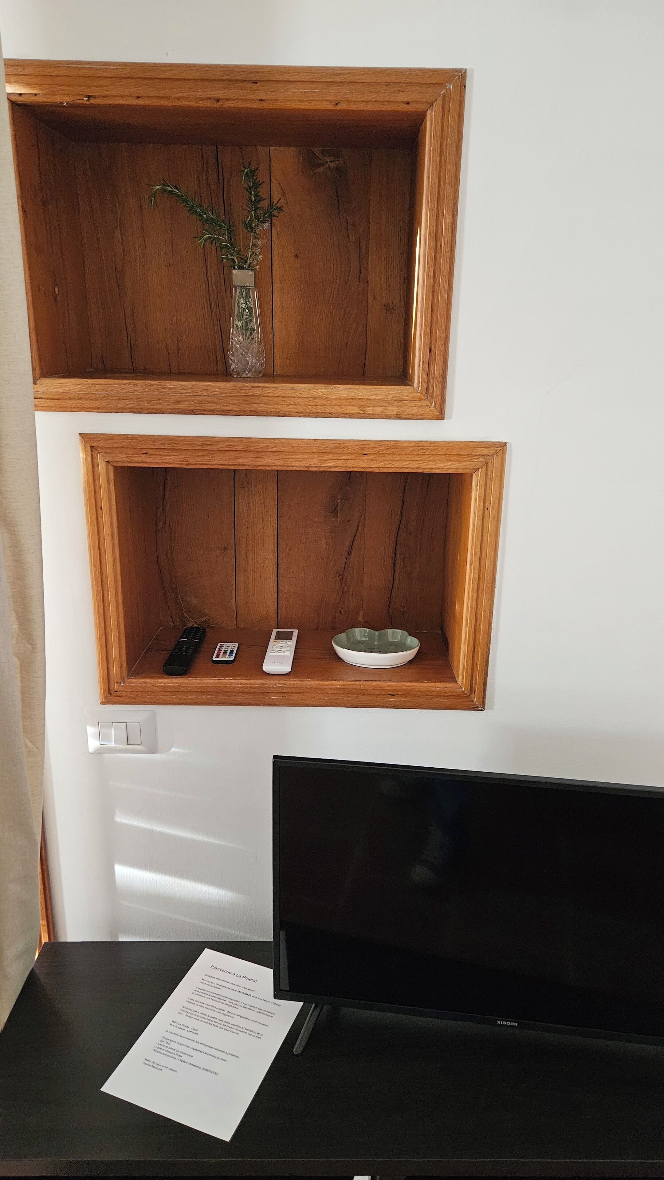 Un estante de madera con un florero con follaje y un control remoto. Abajo, un tazón blanco reposa en un estante inferior. Un televisor negro está sobre un pedestal, y hay un papel sobre la mesa.
