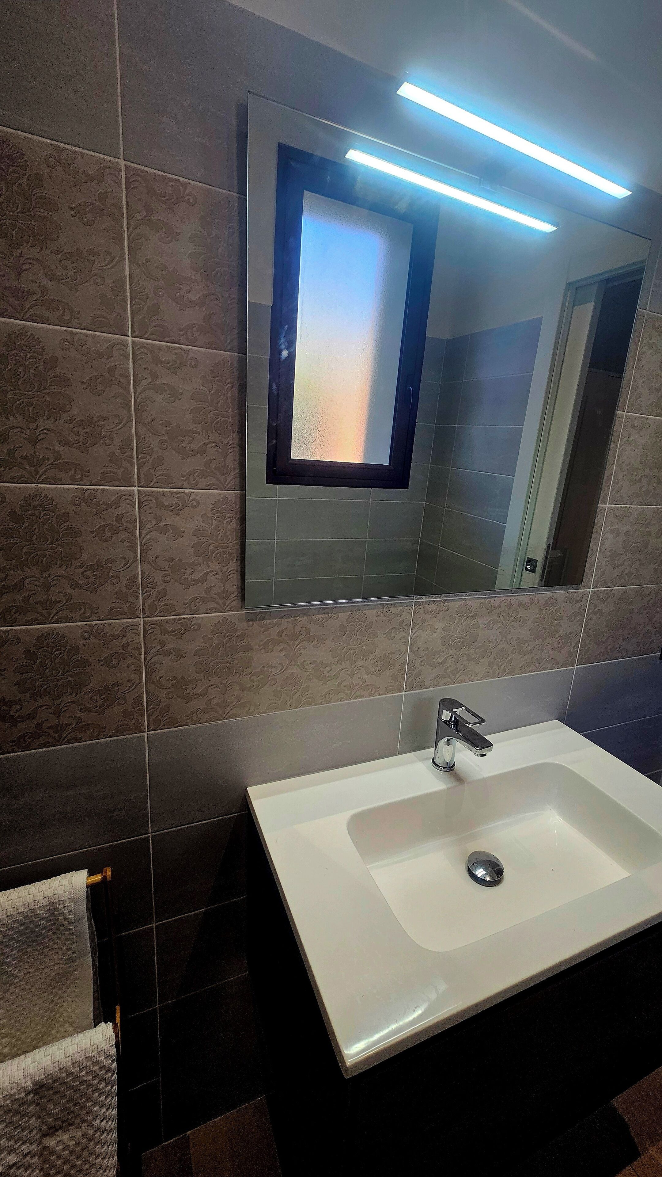 Ein Badezimmer mit einem weißen Waschbecken, dunklen gemusterten Fliesen und einem Spiegel mit satiniertem Glas.