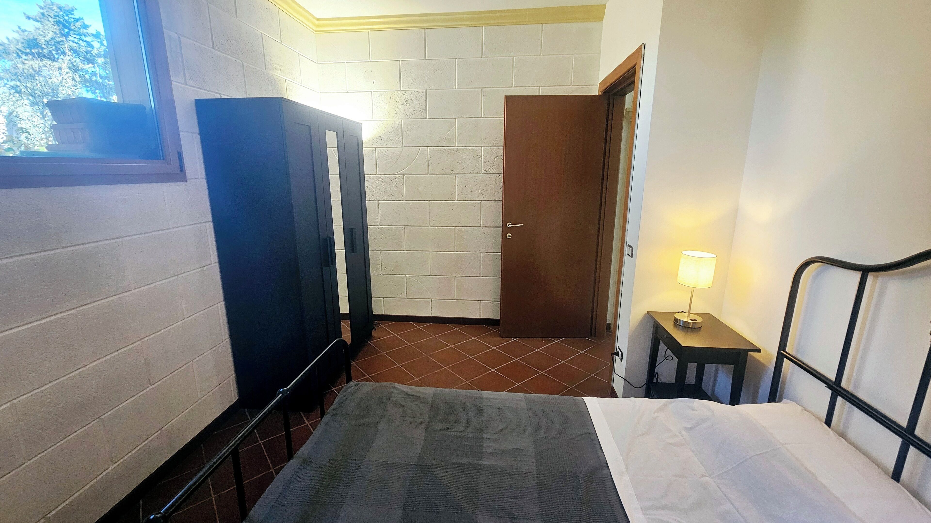 Una habitación pequeña con una cama, una puerta de madera, una mesita con una lámpara y una ventana con vistas a árboles.