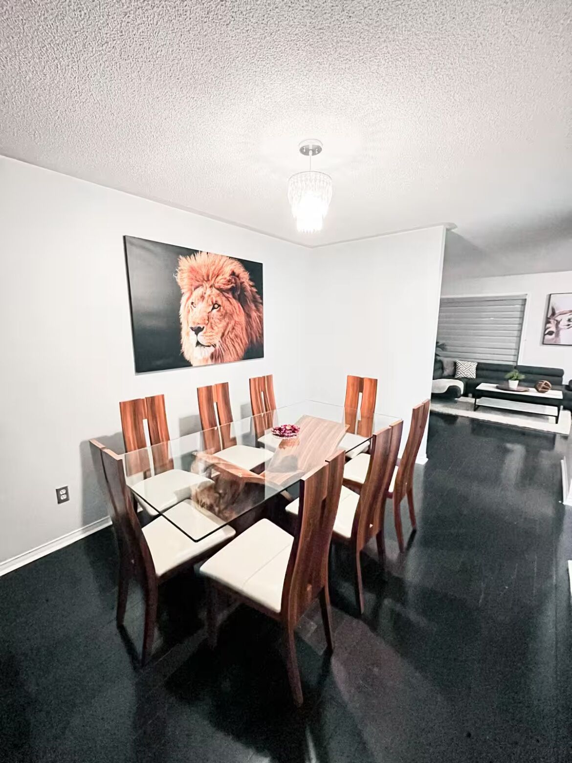 A dining area with a glass-top table and wooden chairs, a large framed picture of a lion on the wall, and a black floor.