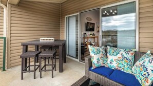A covered patio area with a wicker table and chairs, a sofa with colorful cushions, and a sliding glass door leading to an interior space with a television and decorative items.