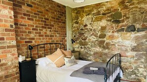 A bedroom with a brick wall, a stone wall, a bed with white bedding, a black nightstand, and a lamp. - Convenient & Cosy Mountain Retreat in Omeo. (Omeo)