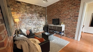 A living room with a stone wall, a black sofa, a television on a wooden stand, and a door to the right. - Convenient & Cosy Mountain Retreat in Omeo. (Omeo)