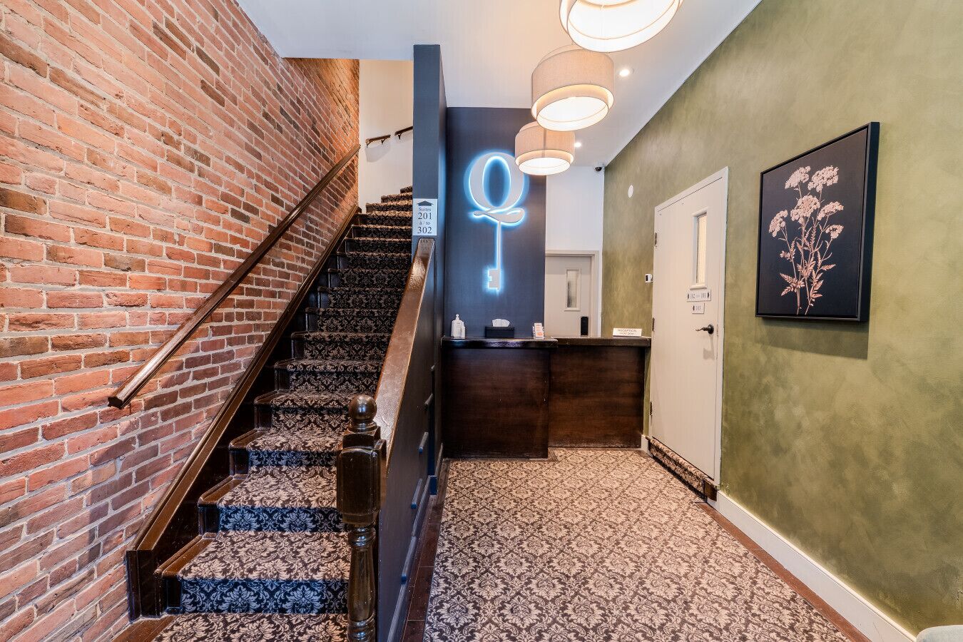 Un escalier recouvert d’une moquette à motifs mène à un espace d’accueil où l’on trouve un panneau lumineux en forme de clé, un comptoir d’accueil et une plante encadrée accrochée au mur.