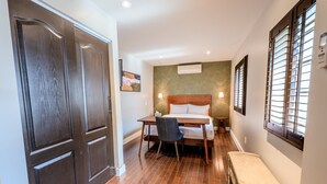 Habitación de hotel con puerta de madera, cama con cabecera de madera, escritorio con silla, mesita de noche con lámpara y ventana con persianas.