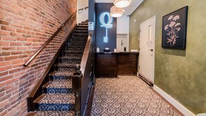 Una escalera con alfombra estampada que conduce a un área de recepción con un letrero de llave de neón, una mesa de recepción y una planta enmarcada en la pared.