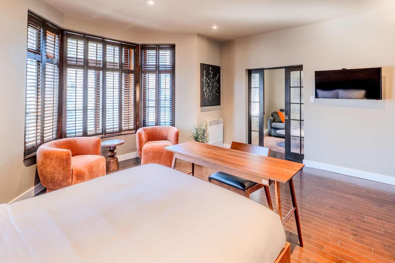 Une chambre d’hôtel avec un grand lit, une table à manger en bois, deux fauteuils et une petite table ronde. De grandes fenêtres sont équipées de stores, un tableau est accroché au mur et un téléviseur est fixé au mur.