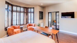 Habitación de hotel con una cama grande, una mesa de comedor de madera, dos sillones y una mesita redonda. Se observan ventanas amplias con persianas, un cuadro en la pared y un televisor montado en la pared.