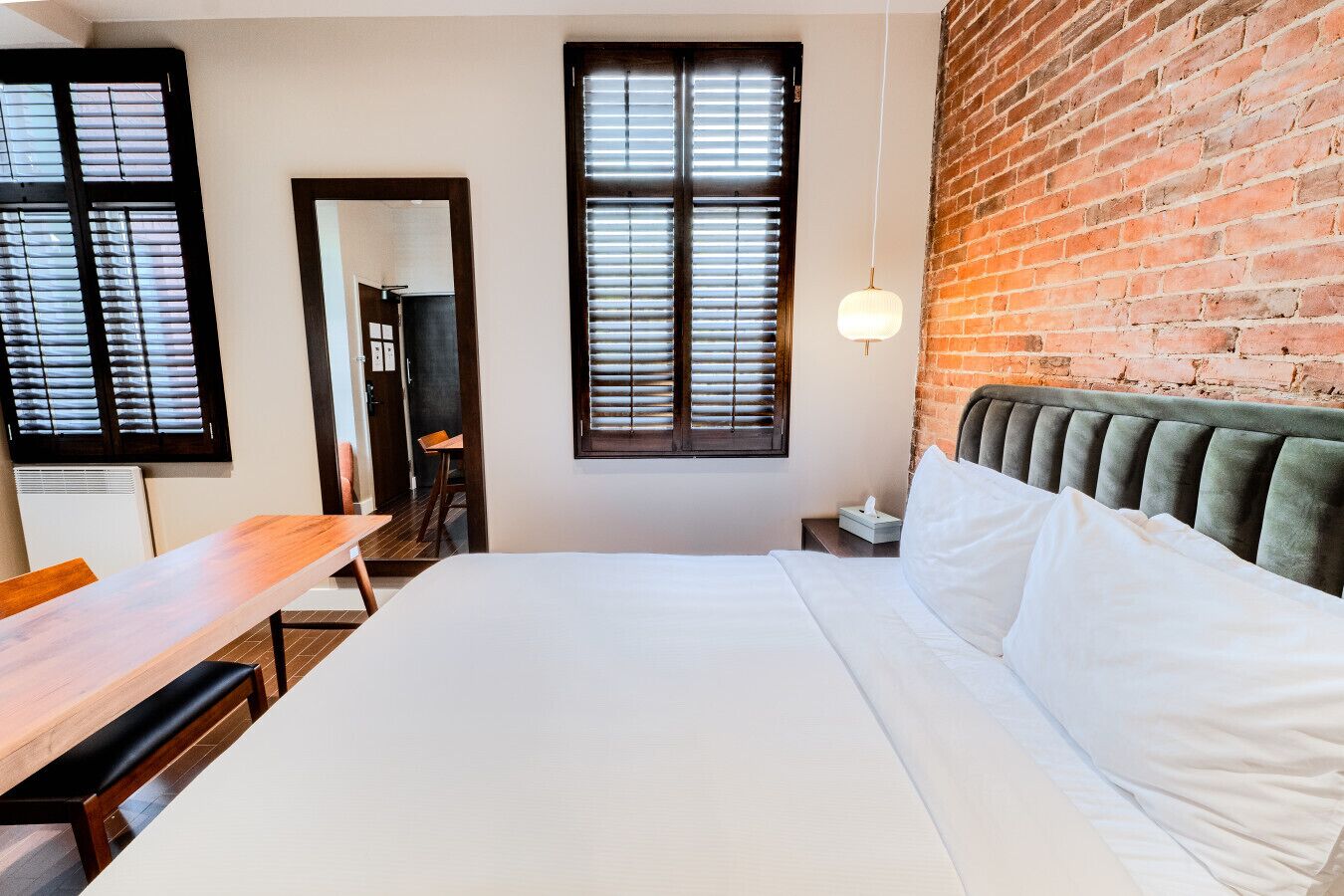 Une chambre d’hôtel avec un lit, un bureau en bois, une chaise, une table de chevet, une lampe et un mur en briques.