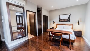 A hotel room with a wooden floor, a bed with white linens, a wooden desk, a chair, a nightstand, and a painting on the wall.