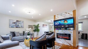 Uma sala de estar moderna com televisão de tela plana, sofá cinza, cadeiras pretas, lareira e um espaço de jantar.