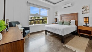 A modern bedroom with a large bed, a desk, and a chair. There is a window with a view of palm trees and a balcony. - Cozy 2BR in Versalles – Walk to Beach & Dining (Puerto Vallarta)