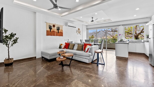 A modern living room with a grey sofa, wooden coffee table, and a kitchen area with white cabinets and a large window. - Cozy 2BR in Versalles – Walk to Beach & Dining (Puerto Vallarta)