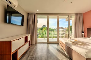 Une chambre d’hôtel avec un grand lit, un téléviseur fixé au mur, une commode en bois et un balcon offrant une vue sur la ville.