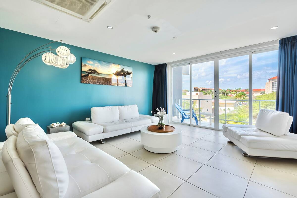 A modern living room with a teal accent wall, white sofas, a round coffee table, and a large window with a view of buildings and greenery.