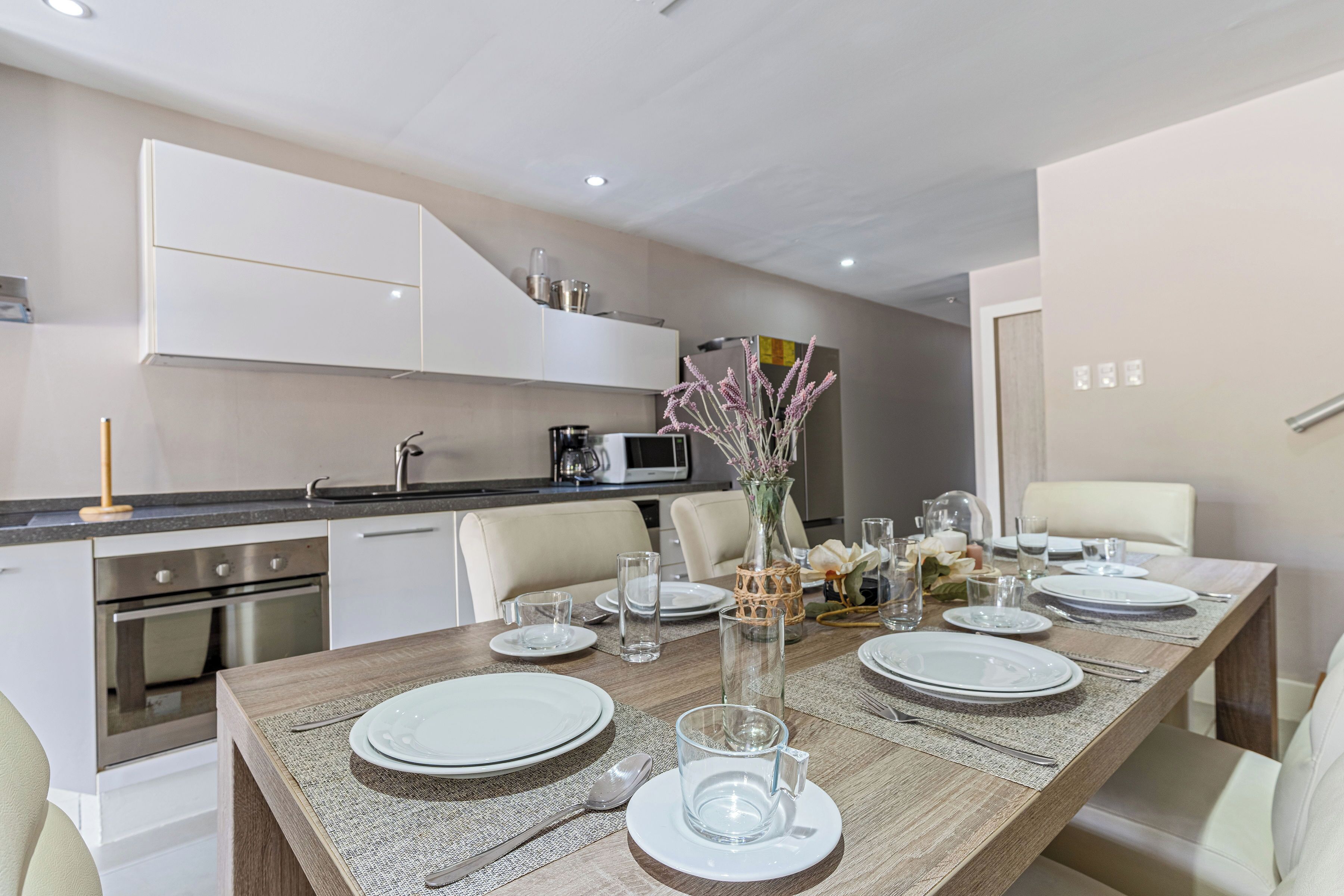A modern kitchen with built-in appliances, a dining area with a wooden table set for four, and a vase with flowers on the table.