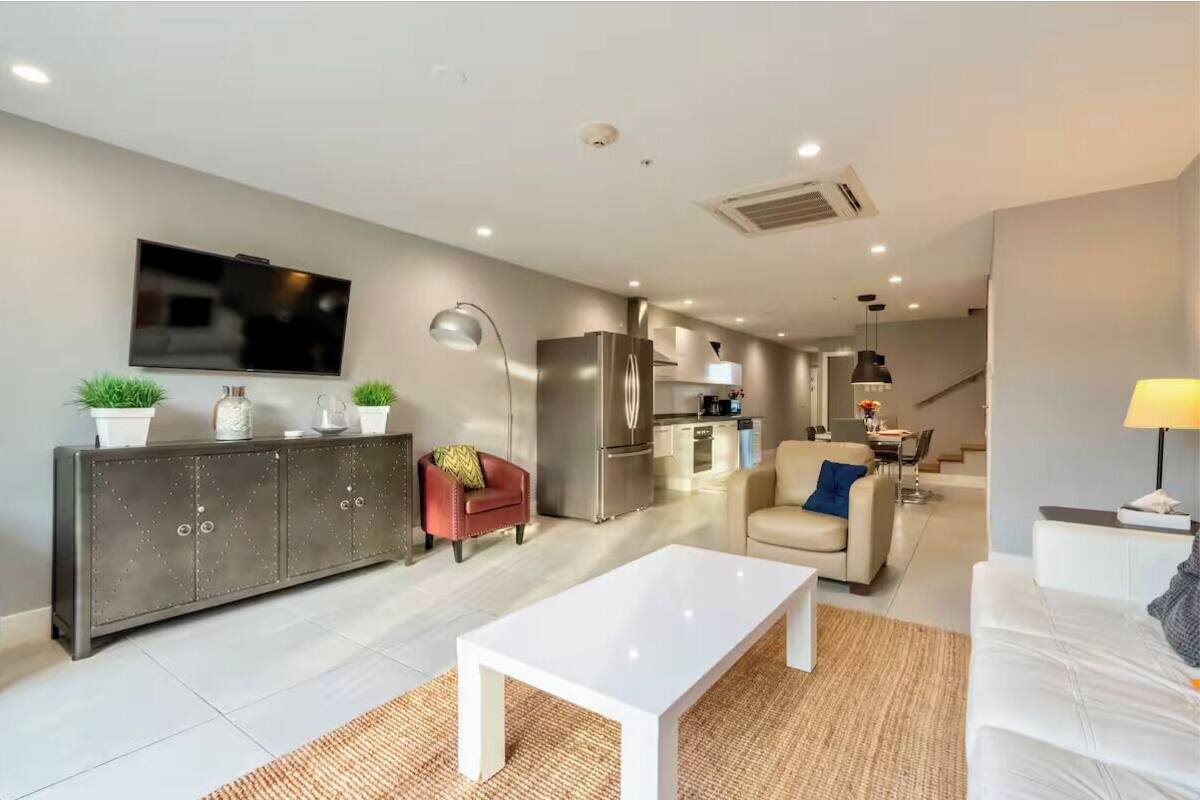A modern living room with a flat-screen TV, a sofa, a coffee table, and a dining area with a kitchen in the background.