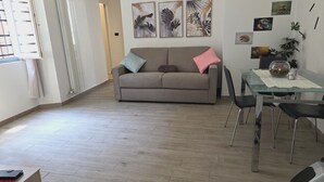 A modern living room with a grey sofa, a glass dining table, and a coffee table. There are wall art pieces and a potted plant on the wall. - Central and quiet apartment, 1 min from the Duomo and 3 min from Lake Como. (Como)