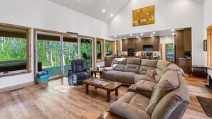A spacious living room with a large sectional sofa, a coffee table, and a dining area with wooden cabinets. Large windows provide a view of the outdoors. - Picturesque Cedar Retreat w/ Hot Tub (McCall)