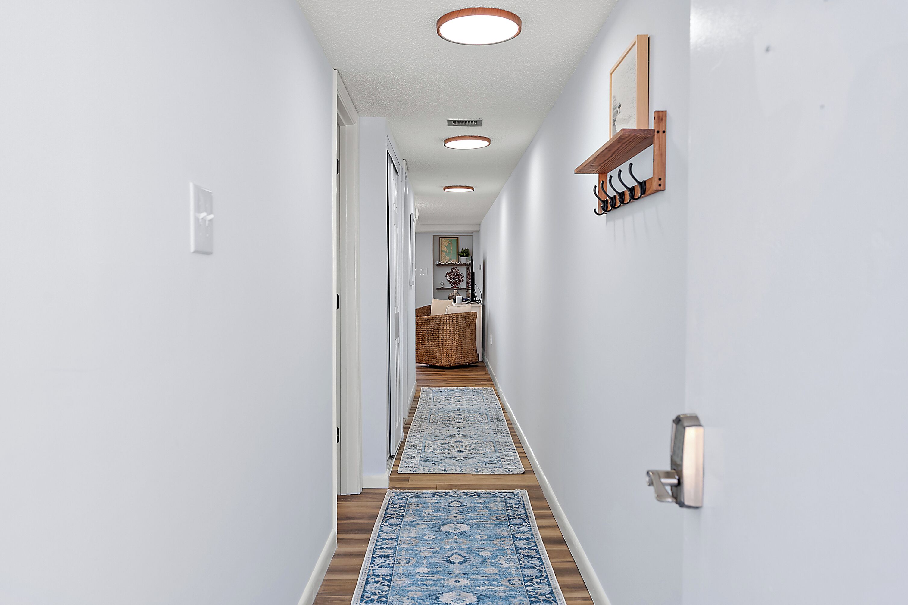 Un pasillo con paredes blancas, suelo de madera y un tapete azul estampado. Se observan tres luces empotradas en el techo y un estante de madera con ganchos en la pared derecha.