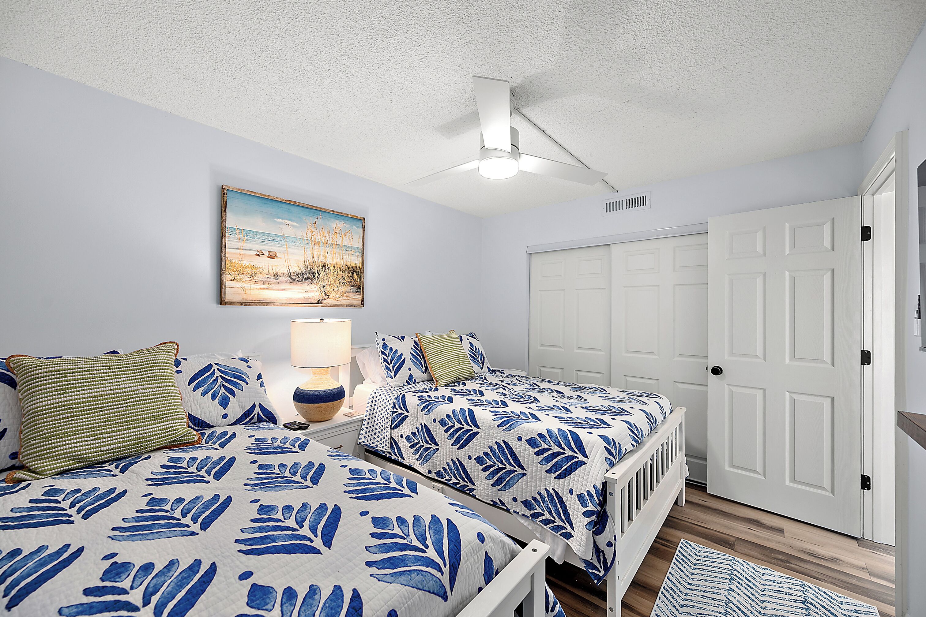 A bedroom with two beds, a ceiling fan, a painting, and a white door.
