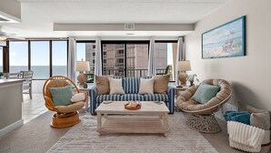 A spacious living room with a large window, a striped sofa, and a beach-themed painting. - Oceanfront 2 Bed/2Bath,IndoorPool,Game Room (Ocean City)
