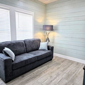 A modern living room with a dark grey sofa, two white pillows, a floor lamp, and a window with blinds.