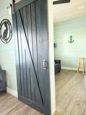 A room with a dark wooden sliding door, a grey sofa, a wooden stool, and a wall-mounted anchor decoration.